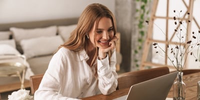 Hymyilevä nainen istuu kotona tietokoneella työpöydän ääressä