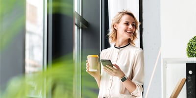 Hymyilevä nainen puhelin ja kahvi kädessä toimistolla