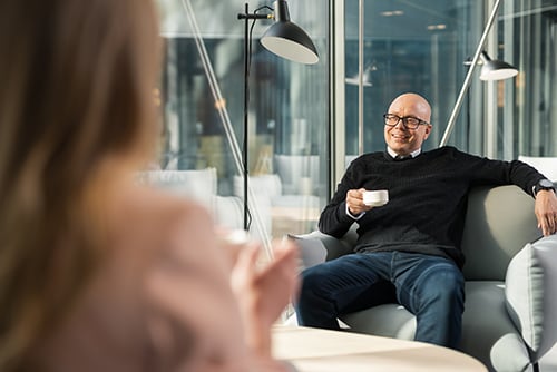 Mies istuu kahvilassa nojatuolissa kahvikuppi kädessään kollegaa vastapäätä.