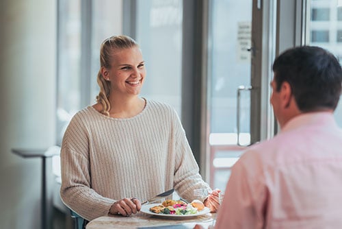 Kollegat syömässä lounasravintolassa. Kuvassa hymyilevä nainen ja mies selkä kohti kameraa.