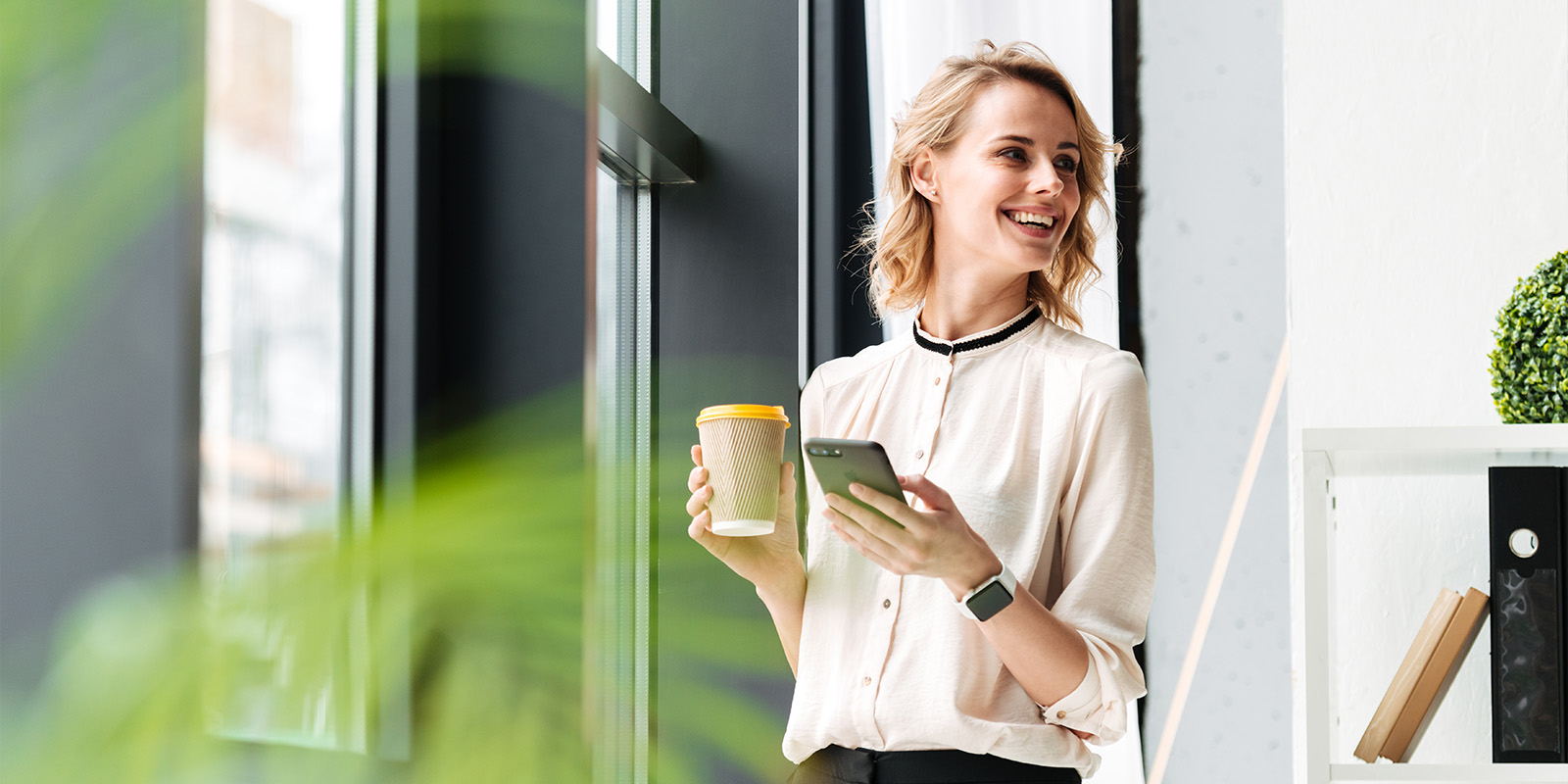 Hymyilevä nainen puhelin ja kahvikuppi kädessä toimistolla