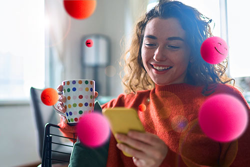 Hymyilevä nainen puhelin ja kahvikuppi kädessä. Ympärillä punaisia ja pinkkejä emojipalloja kuvaamassa Edenredin työsuhde-etujen tuomaa iloa.