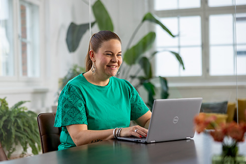 Nainen istuu pöydän ääressä tietokoneella ja työskentelee hymyillen