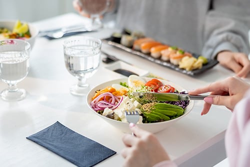 Nainen syö salaattia pöydän ääressä, vieressä istuva ystävä syö sushia