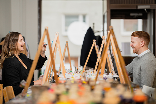 Nainen ja mies maalaamassa tauluja. Taustalla näkyy useita maalaustelineitä.