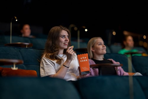 Kaksi naista istuu elokuvateatterissa katsomassa valkokangasta ja syömässä popcornia