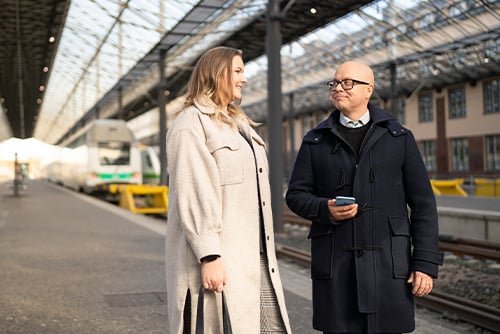 A woman and a man are standing at the train station next to the train tracks and the man has a phone in his hand
